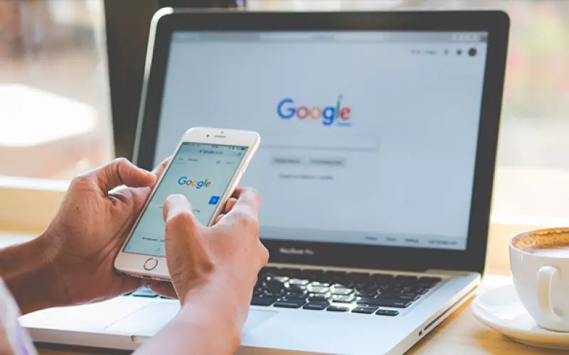 Hands using a phone beside a laptop showing a search page