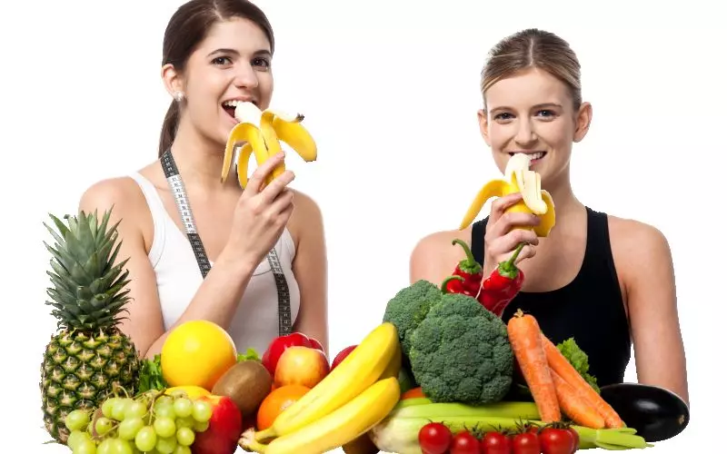 Two women eating bananas beside a pile of fresh fruit and vegetables